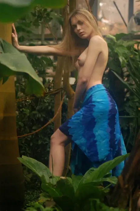 Topless after removing her dress, a blonde woman stands in a lush garden