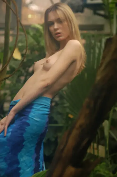 A blonde woman removes her dress and reveals a topless figure in a lush garden