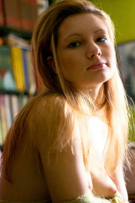 Fully nude, a stunning woman with long legs and a tight rear poses in a library setting