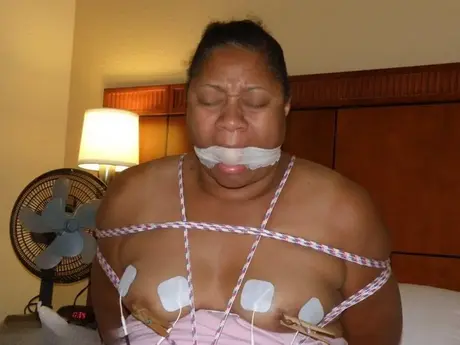 Gefesselte und geknebelte dunkelhäutige BBW erträgt Stromschläge auf einem Bett