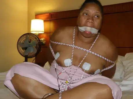 Gefesselte und geknebelte dunkelhäutige BBW erträgt Stromschläge auf einem Bett