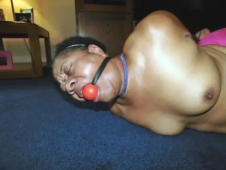 Trixie, une femme noire en surpoids, se débat dans un hogtie tout en portant un bâillon-boule