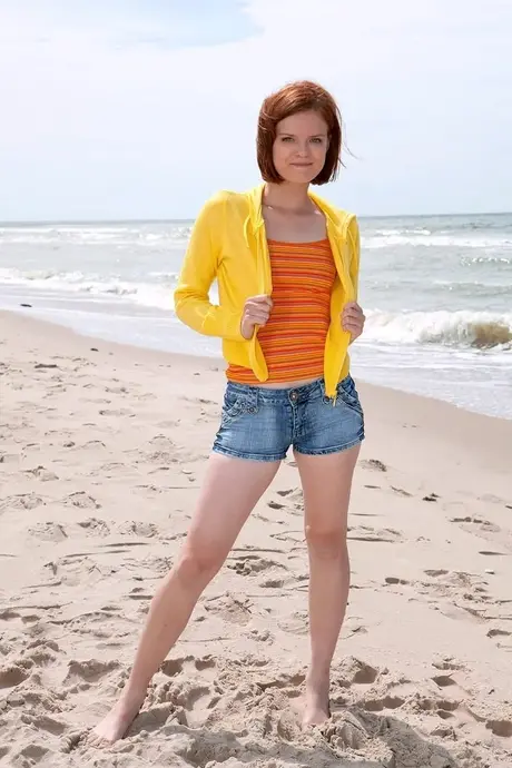 Rudowłosa Denisa o krótkich włosach odsłania swoje nagie ciało na plaży w żółtej bluzie z kapturem